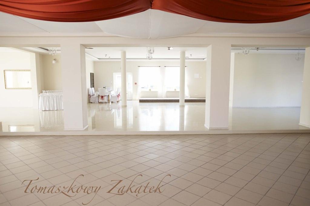 Pusta sala weselna z błyszczącą podłogą w jasnych płytkach, dekoracją z czerwonego materiału na suficie i stołami nakrytymi białymi obrusami w tle.
