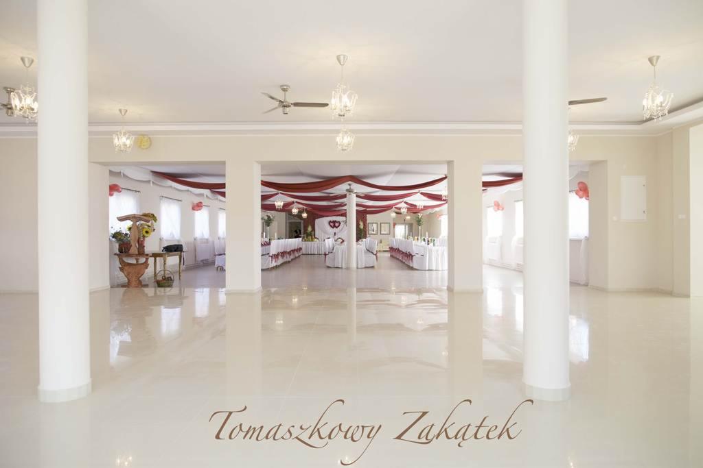 Elegancka sala weselna z kolumnami, dekoracjami z czerwonej tkaniny i białymi obrusami, widok z perspektywy wejścia, błyszcząca podłoga z odbiciem światła.