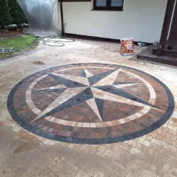 Okrąg z kostki brukowej ułożony w kształt kompasu, z czarną obwódką, na tle jasnej kostki, obok ściany domu i worka z ziemią.