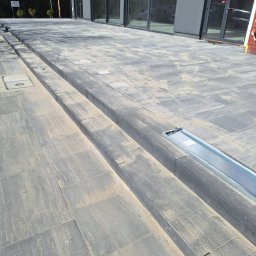 HALINA DANISZEWSKA BRUKARSTWO - Ułożona, szara kostka brukowa z odpływem liniowym przed budynkiem z dużymi oknami. Widoczne schody z kostki. Całość zasypana piaskiem.