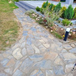 HALINA DANISZEWSKA BRUKARSTWO - Ścieżka z nieregularnych płyt kamiennych prowadząca do domu z nowoczesnym ogrodem. Widoczne rośliny ozdobne i trawnik. Oświetlenie ścieżki w postaci słupków.