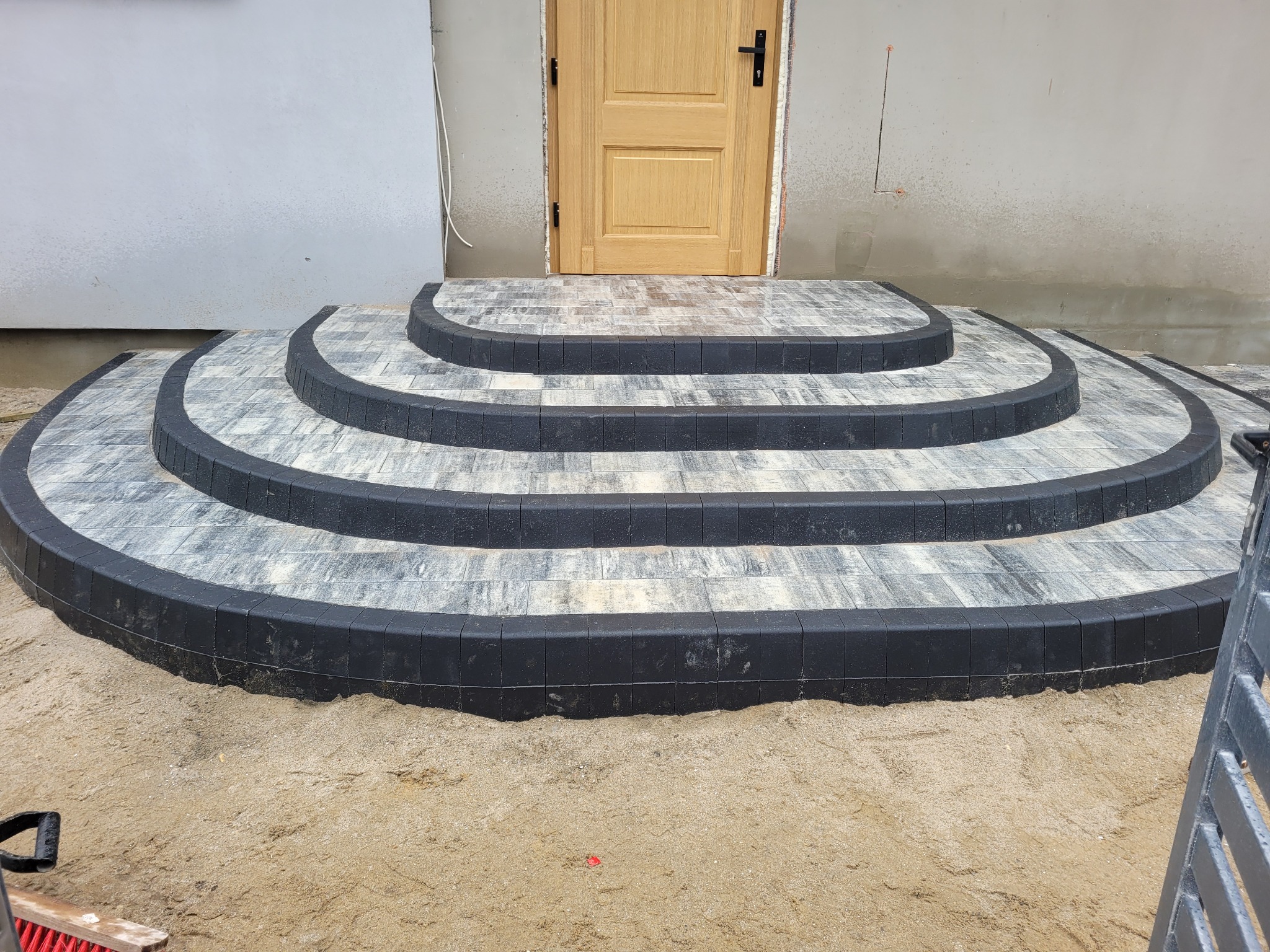 Trzy półokrągłe stopnie wejściowe ułożone z szarej kostki z czarnym obrzeżem, prowadzące do drewnianych drzwi wejściowych, na piasku przed budynkiem.