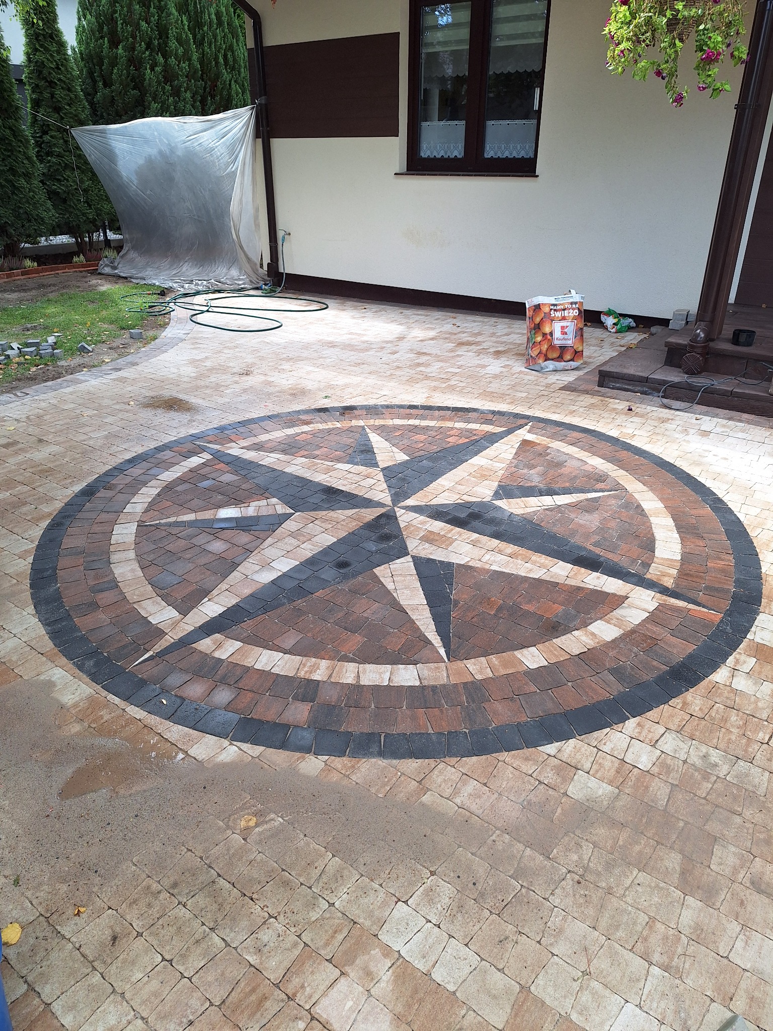 Okrąg z kostki brukowej ułożony w kształt kompasu, z czarną obwódką, na tle jasnej kostki, obok ściany domu i worka z ziemią.