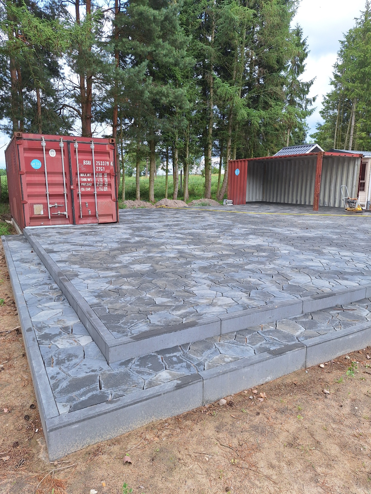 Ułożona kostka brukowa w nieregularnym kształcie, obramowana szarym obrzeżem, z kontenerami w tle. Widok na plac z kostki z dwoma stopniami.