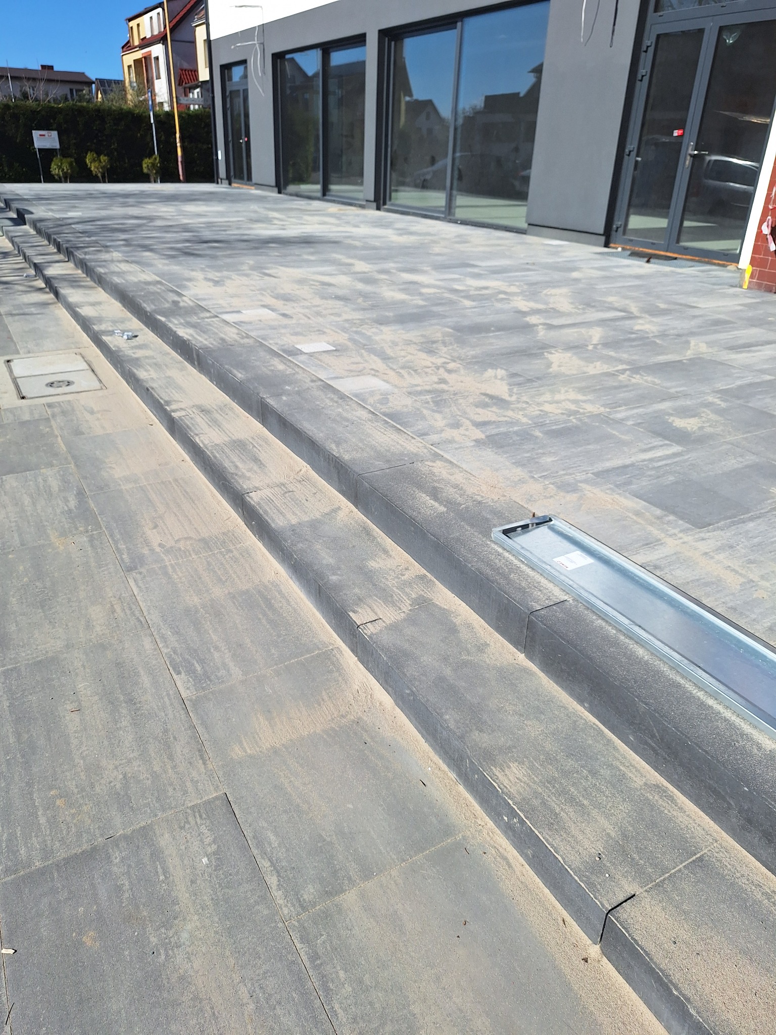 Ułożona, szara kostka brukowa z odpływem liniowym przed budynkiem z dużymi oknami. Widoczne schody z kostki. Całość zasypana piaskiem.