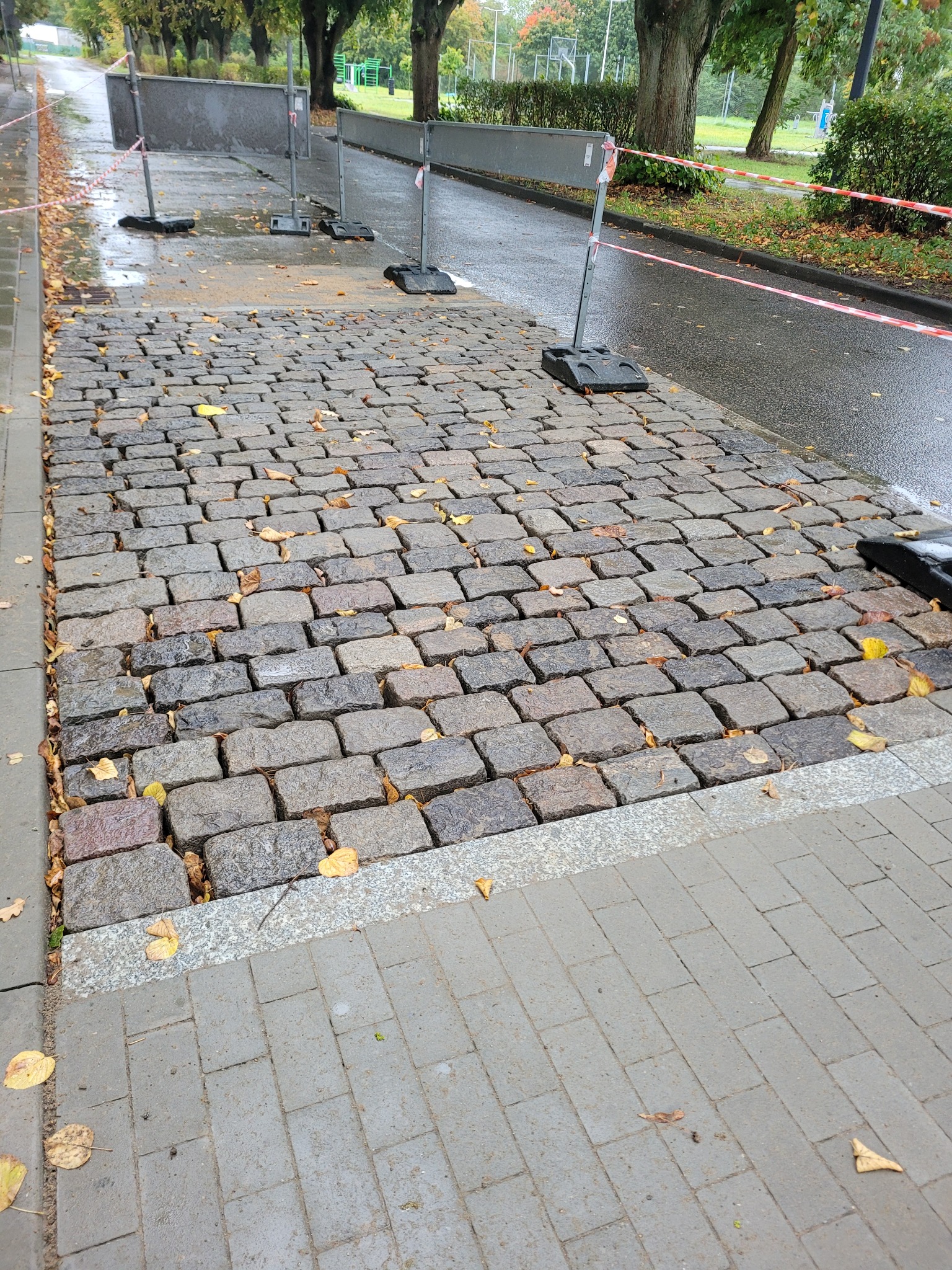 Ułożona kostka granitowa w jesiennej scenerii, zabezpieczona barierkami. Widoczne liście na nawierzchni i mokra powierzchnia po deszczu. Kąt ujęcia z dołu.
