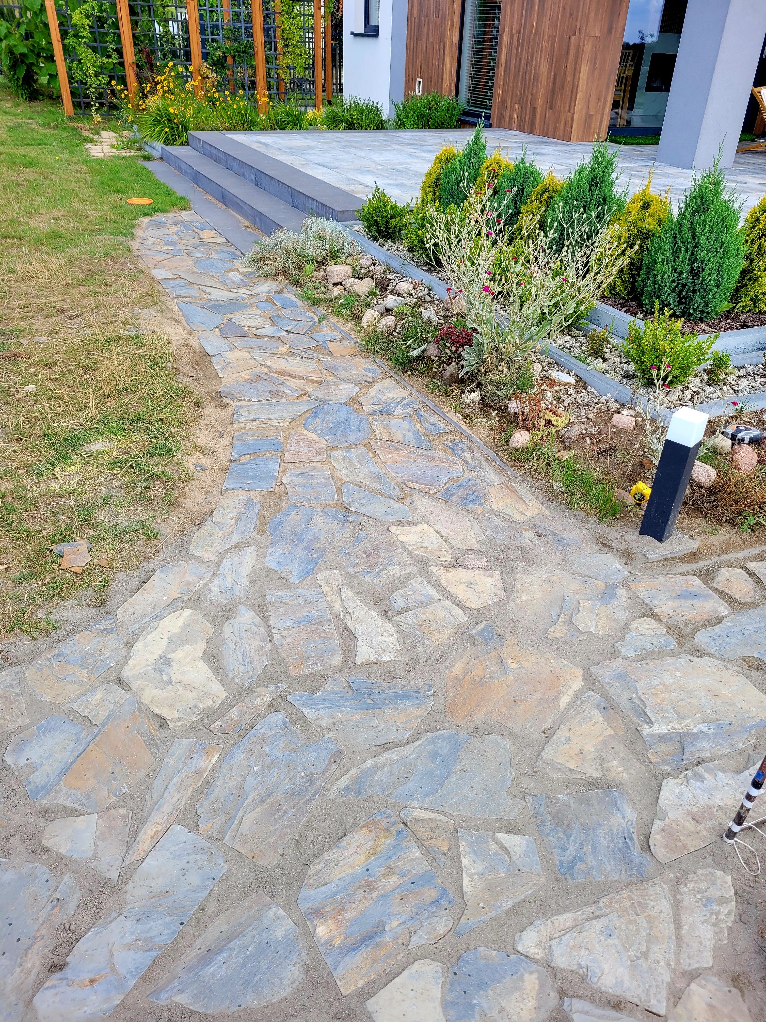 Ścieżka z nieregularnych płyt kamiennych prowadząca do domu z nowoczesnym ogrodem. Widoczne rośliny ozdobne i trawnik. Oświetlenie ścieżki w postaci słupków.