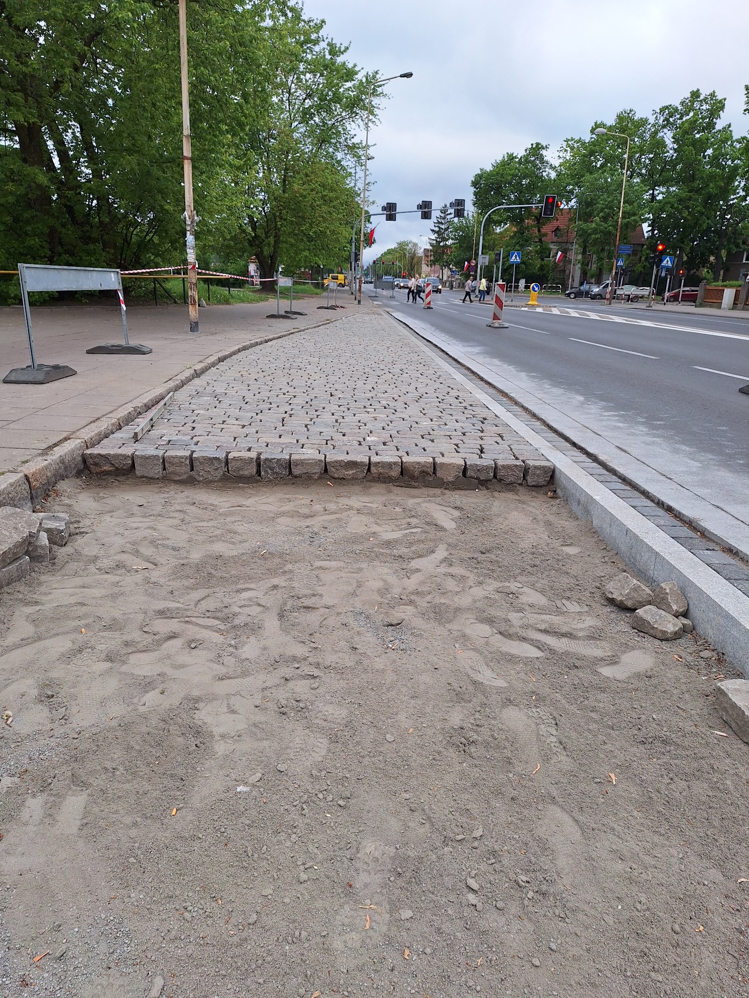 Fragment chodnika z kostki granitowej w trakcie budowy, widoczne przygotowane podłoże i częściowo ułożona nawierzchnia. W tle ulica z sygnalizacją świetlną i przechodniami.