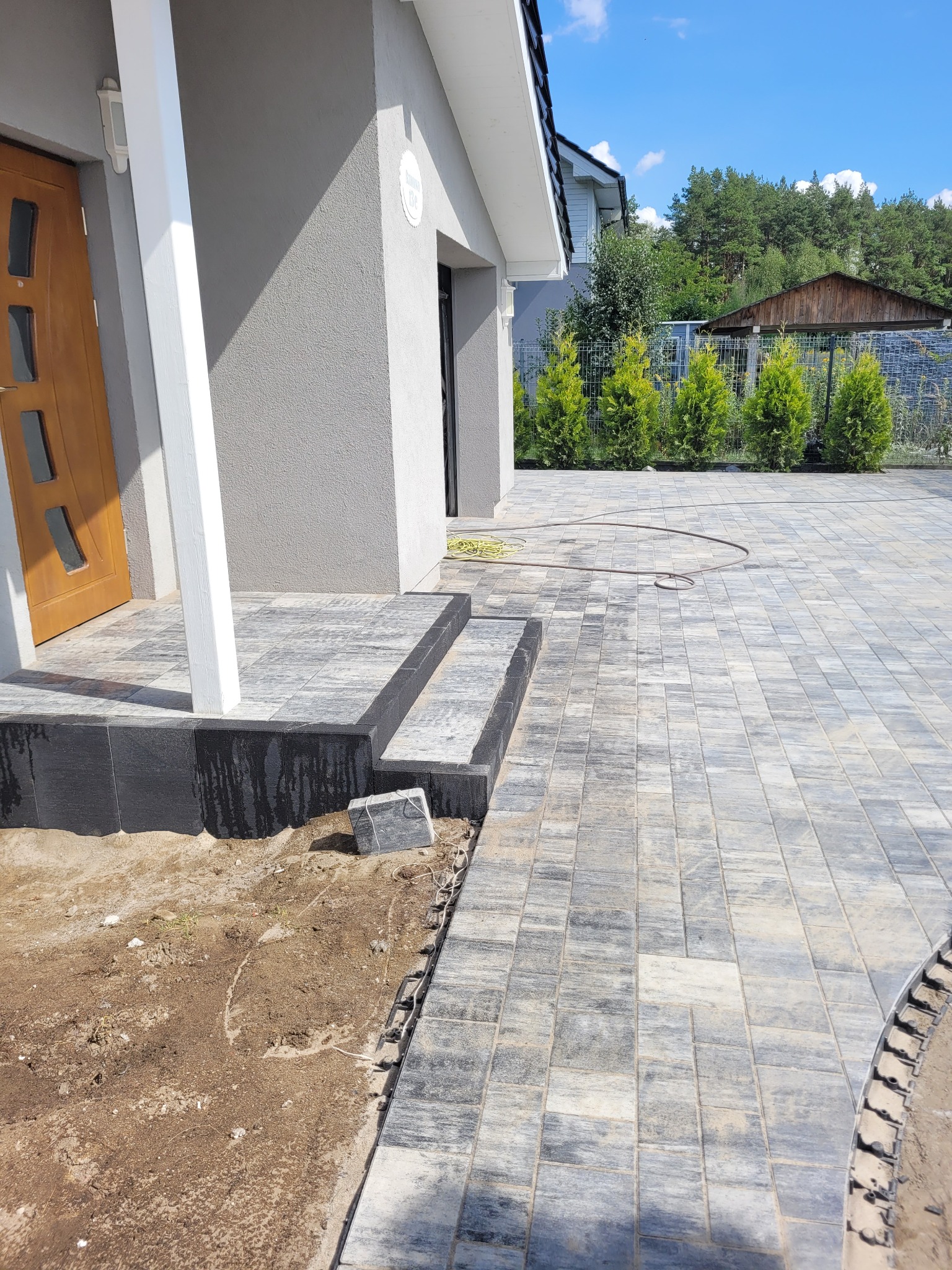 Nowo ułożona, szara kostka brukowa prowadząca do wejścia domu z drewnianymi drzwiami. Widoczne schody z ciemnej kostki i fragment ogrodu z tujami.
