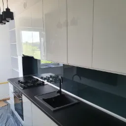 Nowoczesna kuchnia z białymi, lakierowanymi szafkami górnymi i czarnym blatem, widoczny fragment płyty gazowej i zlewu, podłoga zabezpieczona folią malarską.