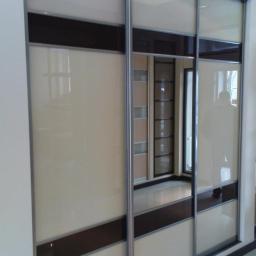 Arkadiusz Nowak - Trzydrzwiowa szafa przesuwna z beżowymi i brązowymi panelami, srebrne ramy, widoczne odbicie wnętrza z półkami.