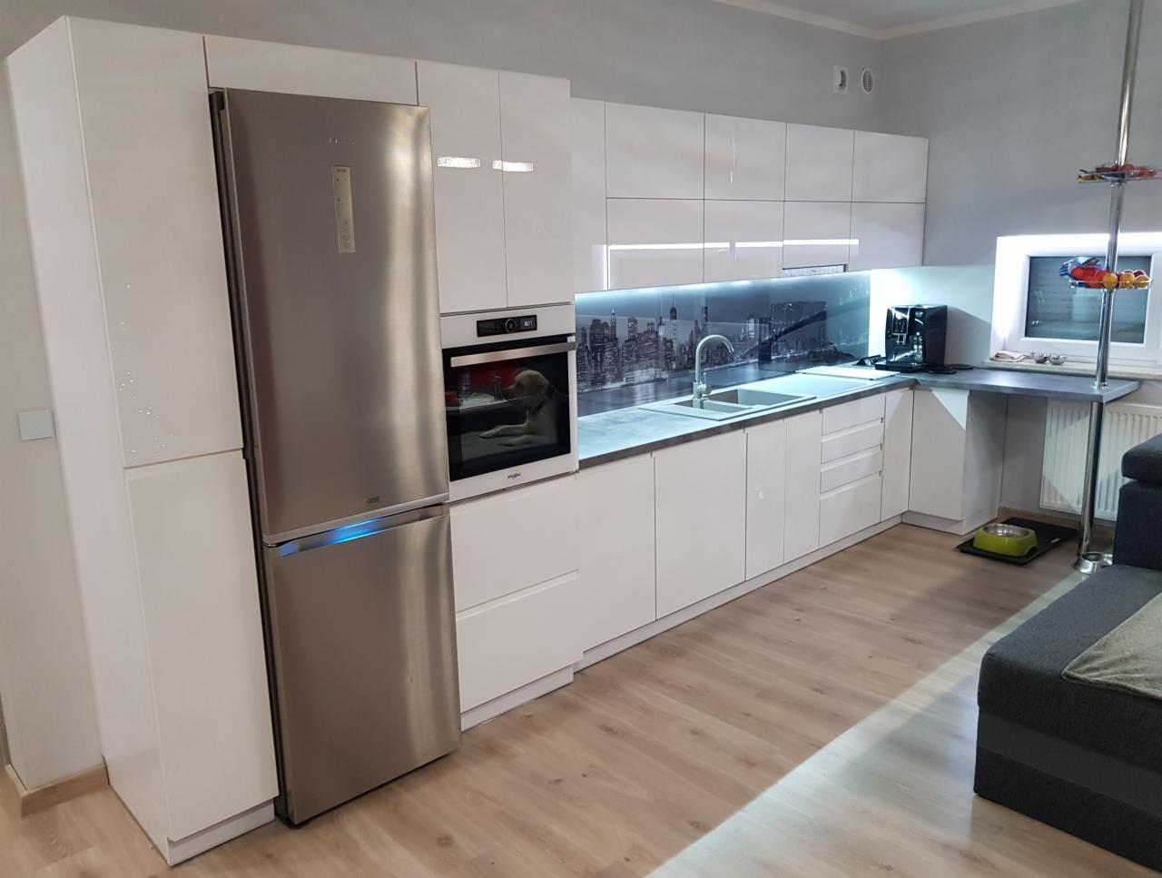 Nowoczesna kuchnia z białymi, błyszczącymi frontami, srebrną lodówką side-by-side, szklanym panelem z motywem panoramy miasta nad blatem i podświetleniem LED.