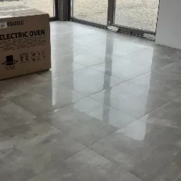 Nowe, szare płytki podłogowe z połyskiem, duże okno z widokiem na pole i karton z napisem ELECTRIC OVEN w nowym domu.