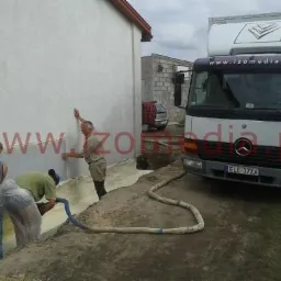 Pracownicy aplikują izolację natryskową na ścianę budynku, widoczny wąż doprowadzający materiał i samochód dostawczy z logo firmy.