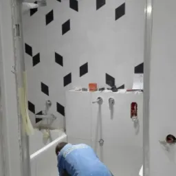 Glazurnik w trakcie układania białych płytek podłogowych w łazience z wzorem w czarne romby na ścianach, widoczne narzędzia i niedokończone prace przy futrynie.