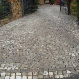 Ułożona z kostki granitowej droga dojazdowa do posesji, wzór wachlarzowy, brama wjazdowa z kutego żelaza w tle.