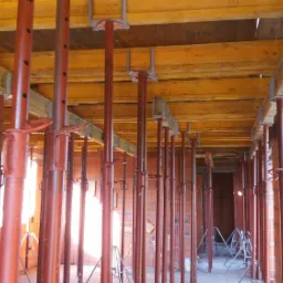 Wnętrze powstającego domu z czerwonej cegły, podpartego licznymi, czerwonymi stemplami budowlanymi, podtrzymującymi drewniany strop.