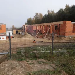 Wznoszenie ścian z czerwonej cegły na budowie domu jednorodzinnego w trakcie wczesnej jesieni, widoczne drewniane nadproża i betonowa mieszarka, ogrodzenie z siatki w pierwszym planie.