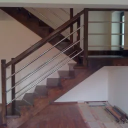 Drewniane schody z metalową balustradą w nowoczesnym wnętrzu. Widoczne elementy wykończeniowe na podłodze, sugerujące trwający montaż.