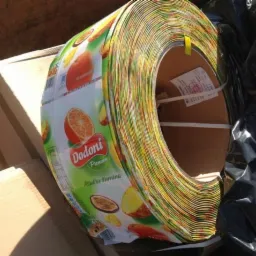 Rolka odpadu folii PVC z nadrukiem owoców (pomarańcze, marakuja, ananas) marki Dodoni, umieszczona w kontenerze na gruz, widoczne kartony i czarny worek na śmieci.