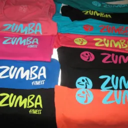 Stos kolorowych koszulek z nadrukiem 'Zumba Fitness', ułożonych w nierównych rzędach, z widocznymi różnymi kolorami nadruków i materiałów.