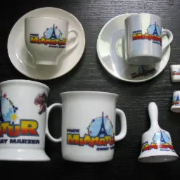 Zestaw pamiątek z Parku Miniatur: filiżanki, kubki, dzwonek i małe pojemniczki z logo parku, ustawione na czarnym tle.
