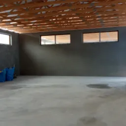 Wnętrze budynku po nałożeniu szarego tynku maszynowego na ściany, widoczne drewniane elementy konstrukcji dachu, dwa worki z zaprawą w rogu pomieszczenia.