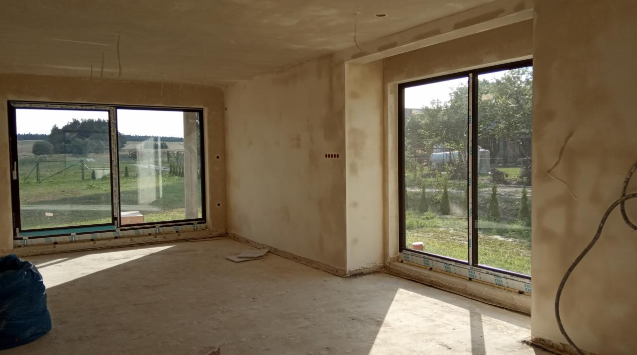Wnętrze domu w trakcie prac wykończeniowych, ściany po nałożeniu szarego tynku, duże okna przesuwne z widokiem na zieleń, na podłodze pozostałości po pracach.