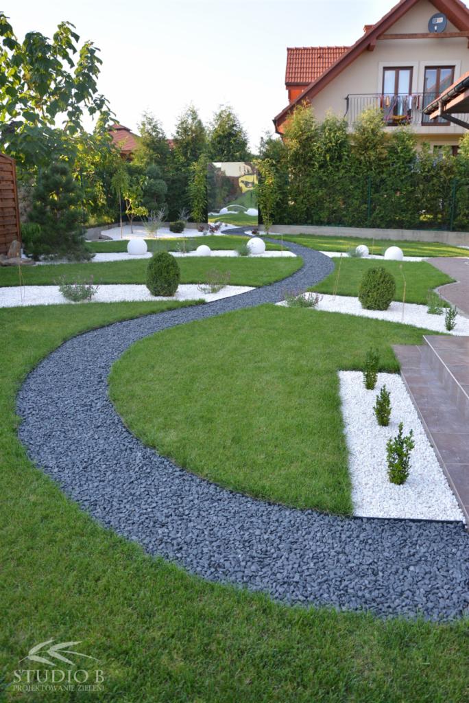 Ogród po metamorfozie: kręta ścieżka z szarego żwiru, trawnik, białe kamienie, kuliste krzewy i oświetlenie. Dom z czerwonym dachem w tle.