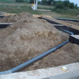 Fundamenty z izolacją i ułożonymi szarymi rurami kanalizacyjnymi w wykopie ziemnym. Widoczna puszka z preparatem uszczelniającym.