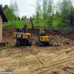 Dwie żółte koparki (Caterpillar i Wacker Neuson) pracują przy wykopie ziemnym na nierównym terenie, w tle budynek i drzewa.