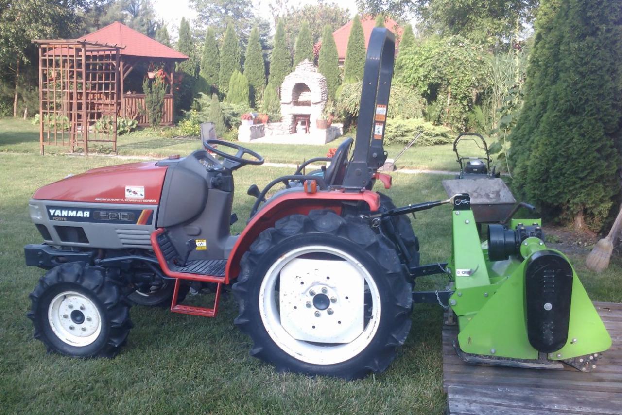 Czerwono-szary mini traktorek Yanmar E210 z podłączonym zielonym frezem glebogryzarki na tle zadbanego ogrodu z altaną i kamiennym grillem.
