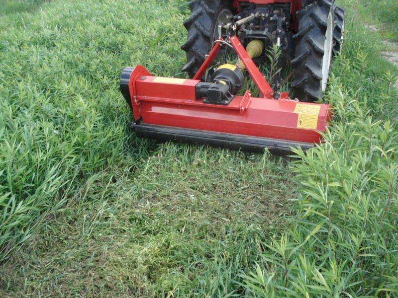 Czerwony mini traktorek z kosiarką bijakową w trakcie pracy na zarośniętej łące, widoczne ślady świeżo skoszonej trawy.