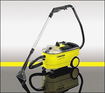 Żółty odkurzacz piorący Karcher Puzzi 100 z przezroczystym zbiornikiem na wodę, widoczny na szarym tle z żółtym akcentem, ukazujący odbicie urządzenia.