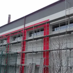 Budynek w trakcie prac elewacyjnych, widoczne rusztowania i fragment pomalowanej na czerwono fasady, pracownik w odblaskowej kamizelce na rusztowaniu.