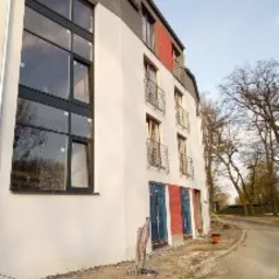 Boczny widok na fragment budynku po nałożeniu jasnego tynku z widocznymi oknami w czarnych ramach, niektóre fragmenty elewacji w kolorze czerwonym, oraz zabezpieczone folią drzwi wejściowe...