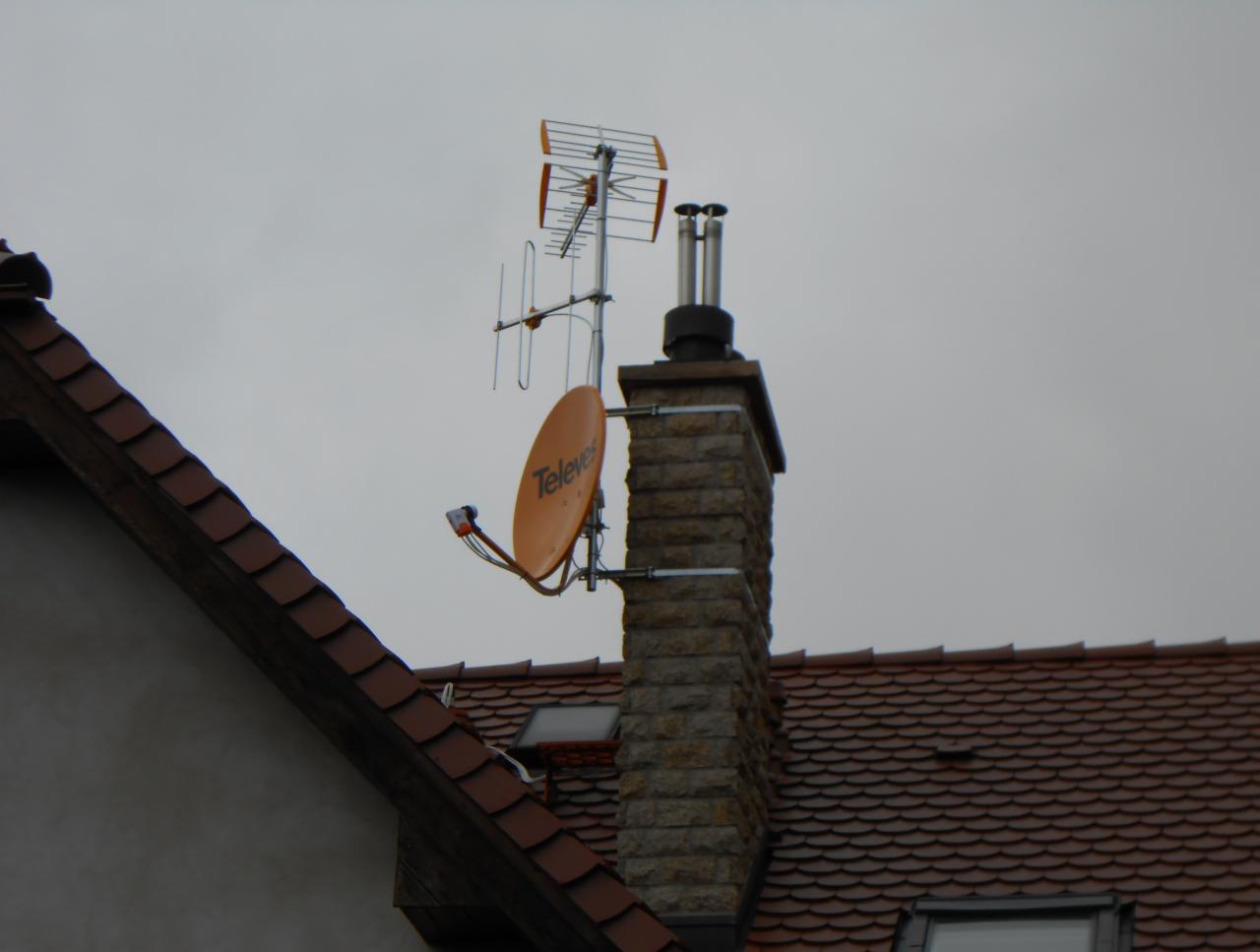 Antena satelitarna Televes i antena naziemna zamontowane na kominie z cegły na dachu budynku z czerwoną dachówką, pochmurne niebo w tle.