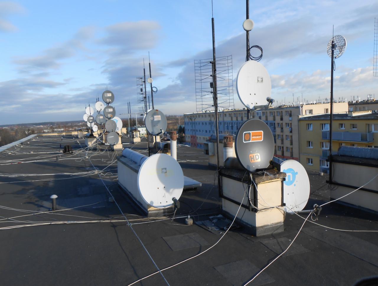 Rząd anten satelitarnych i naziemnych na płaskim dachu budynku wielorodzinnego, widoczne kable i uchwyty, w tle zabudowa miejska i błękitne niebo z chmurami.
