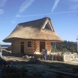 PHU KALBOT s.c. - Budowa domu z bali z dachem krytym strzechą w trakcie prac wykończeniowych, widoczni pracownicy i elementy konstrukcyjne, w tle las i turbina wiatrowa.