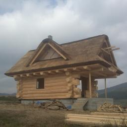 PHU KALBOT s.c. - Surowy dom z bali z dachem krytym strzechą w trakcie budowy, widoczne materiały budowlane i góry w tle.