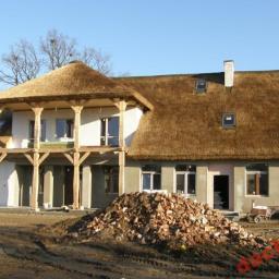 PHU KALBOT s.c. - Dom w trakcie budowy z dachem krytym strzechą, widoczne rusztowania i sterta gruzu na pierwszym planie.