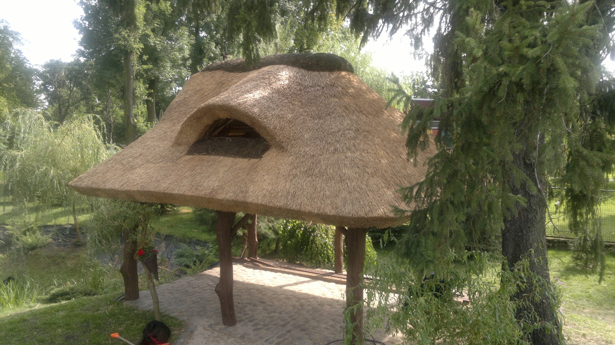 Altana z dachem krytym strzechą, z małym oknem w dachu, wsparta na drewnianych słupach, otoczona zielenią i drzewami.