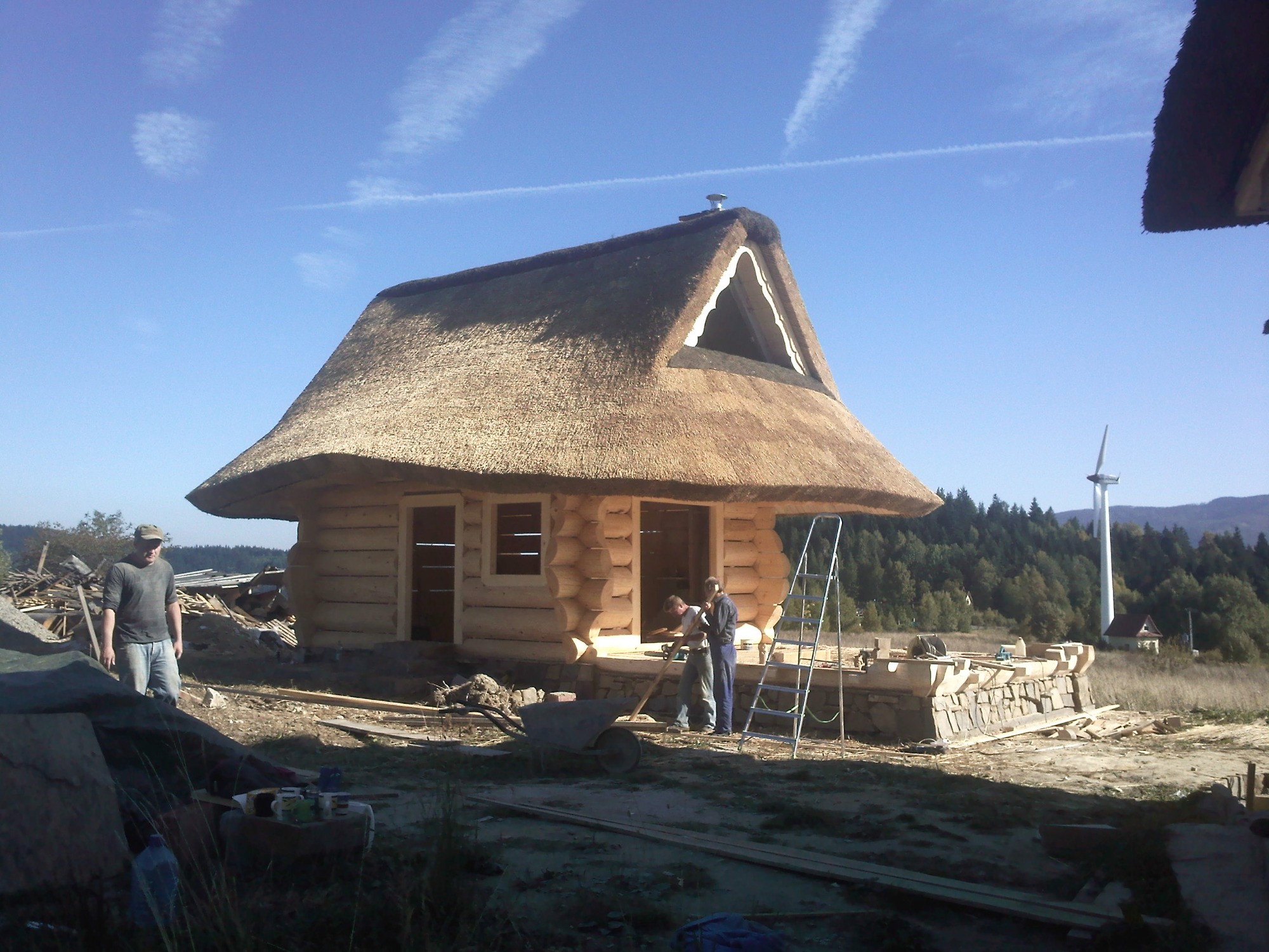 Budowa domu z bali z dachem krytym strzechą w trakcie prac wykończeniowych, widoczni pracownicy i elementy konstrukcyjne, w tle las i turbina wiatrowa.