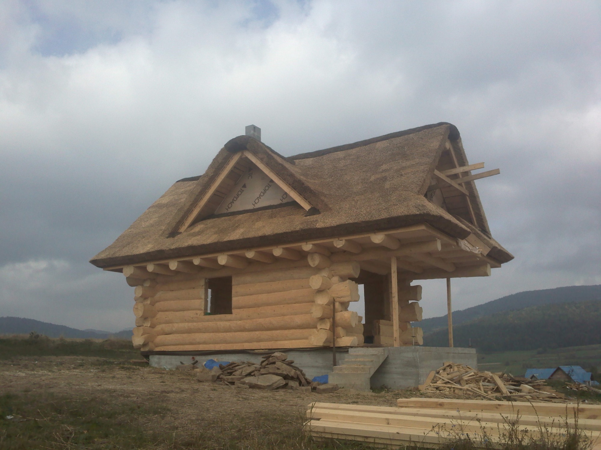 Surowy dom z bali z dachem krytym strzechą w trakcie budowy, widoczne materiały budowlane i góry w tle.