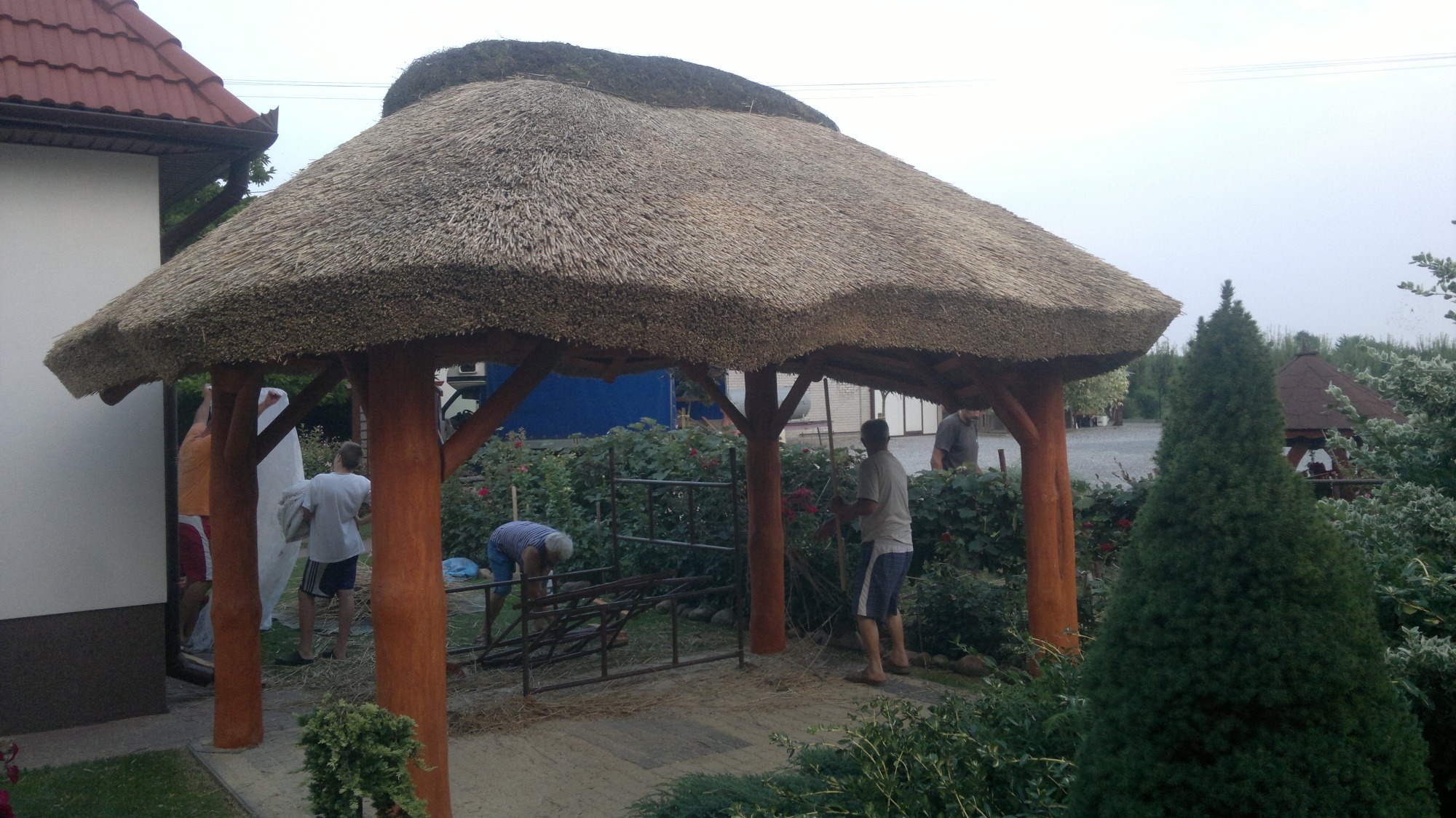 Budowa altany z dachem krytym strzechą, widoczne drewniane słupy konstrukcyjne oraz ekipa montująca elementy wykończeniowe.