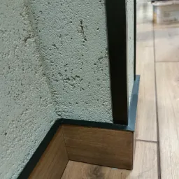 Detal narożnika ściany z dekoracyjną strukturą, drewnianej listwy przypodłogowej z czarnym akcentem i fragmentu drewnianej podłogi.