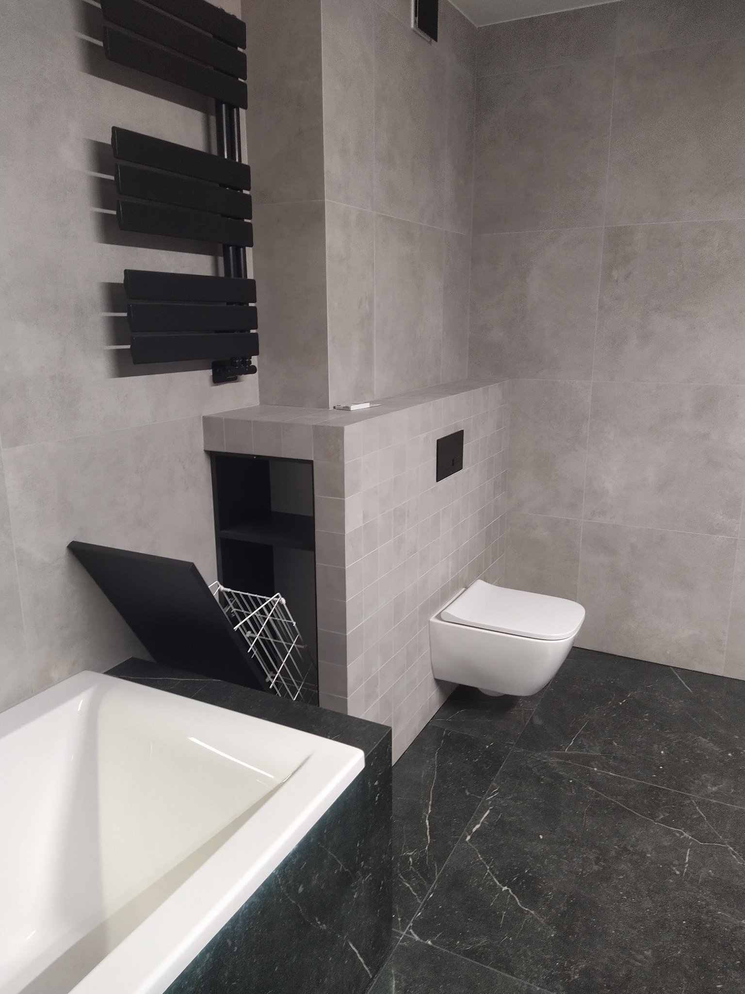 Nowoczesna łazienka z szarymi płytkami, czarnym grzejnikiem drabinkowym i czarną podłogą imitującą marmur, wisząca miska WC i zabudowana wanna z koszem na pranie.