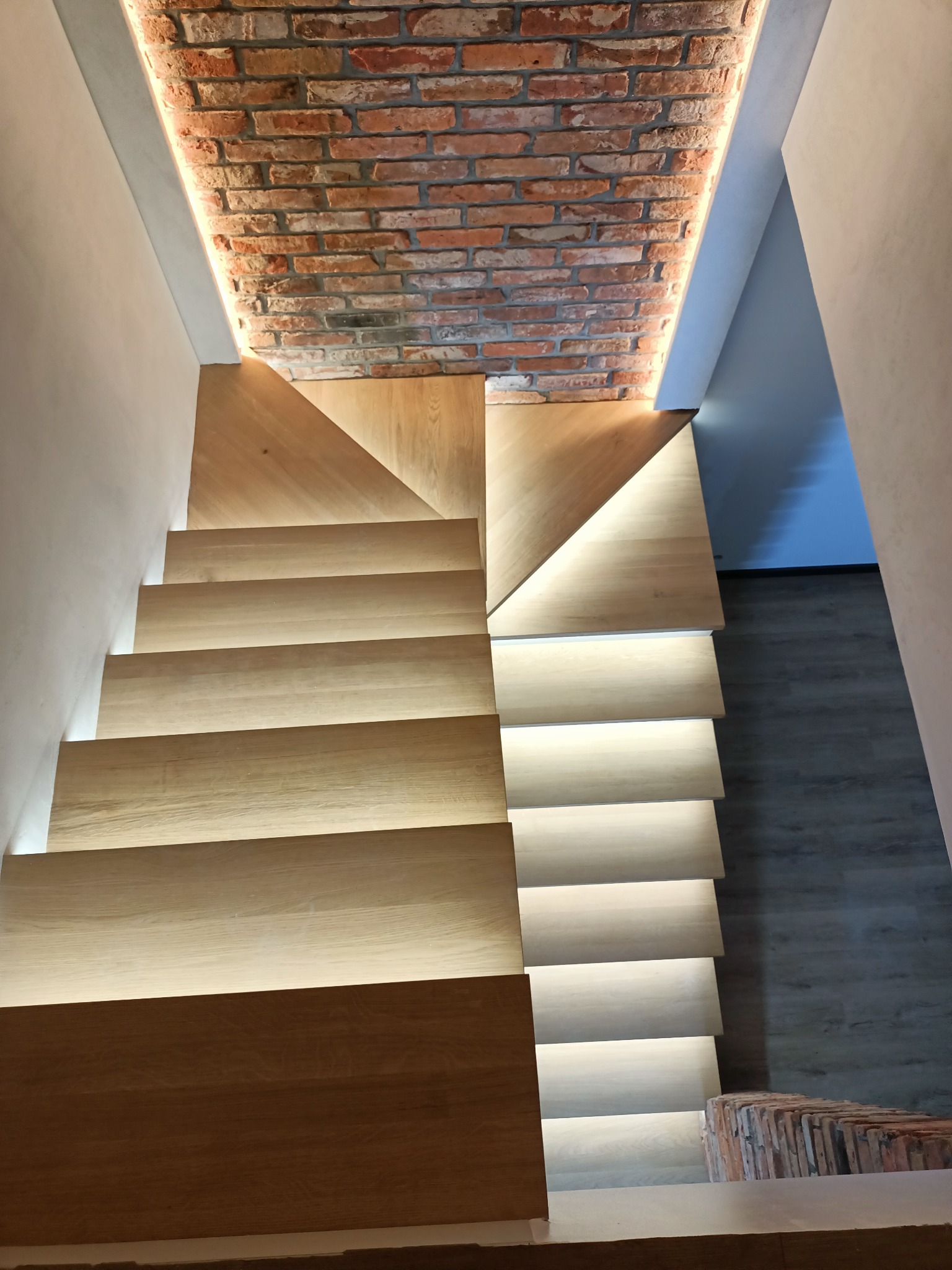 Nowoczesne schody drewniane z podświetleniem LED stopni oraz fragmentu sufitu wykończonego czerwoną cegłą.