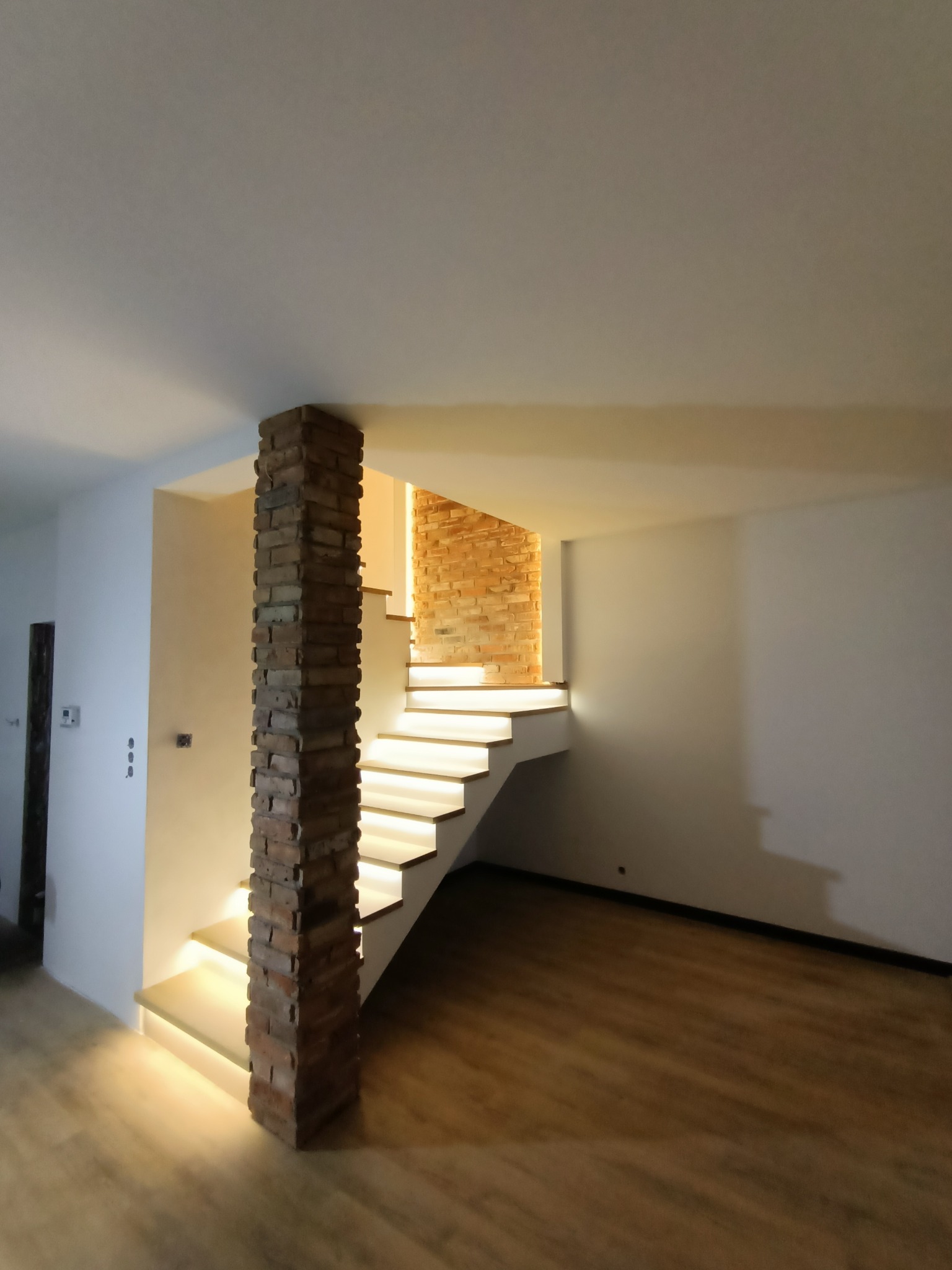 Nowoczesne wnętrze z drewnianą podłogą, białymi ścianami i schodami z podświetleniem LED, wsparte kolumną z cegły.