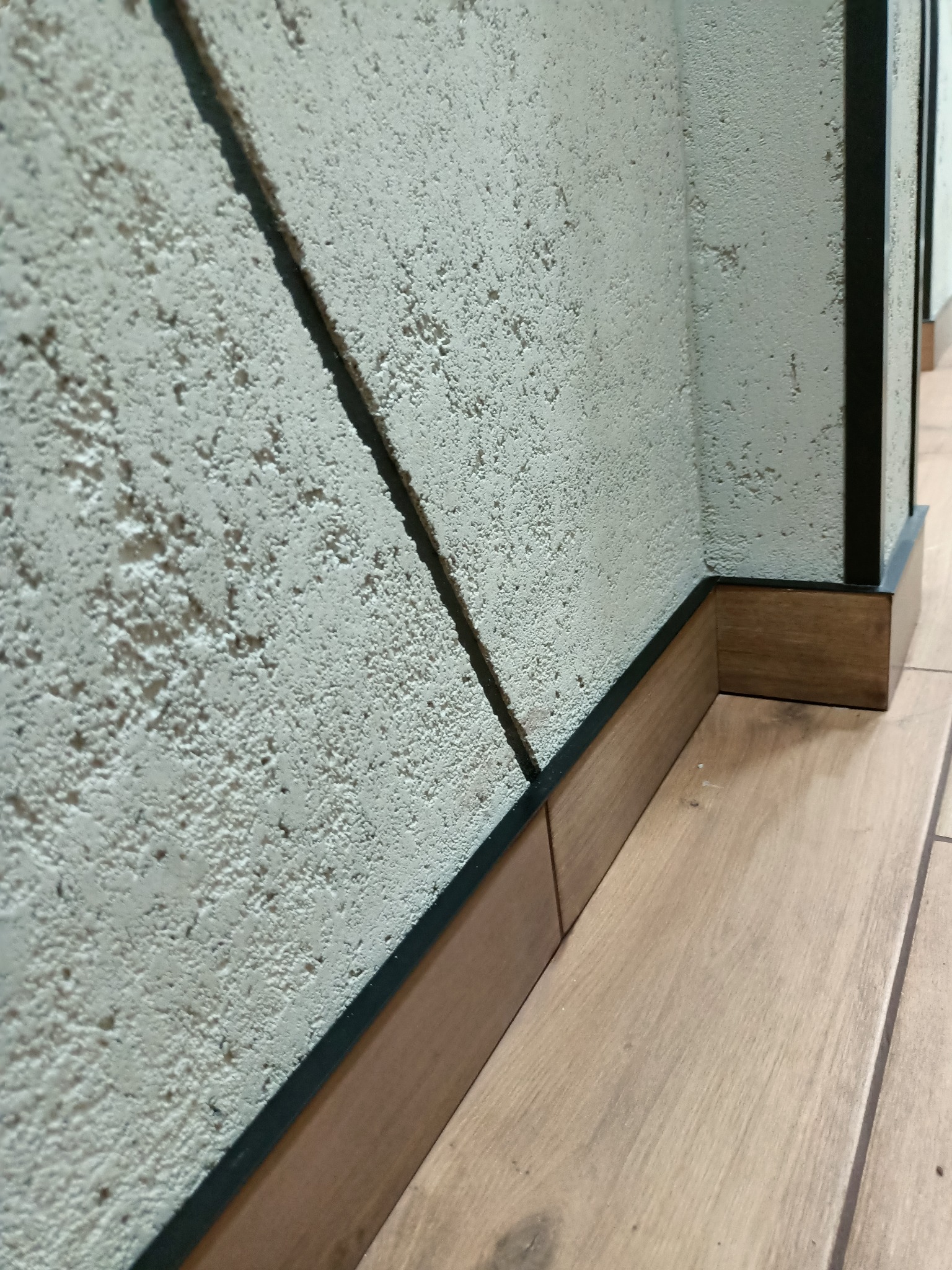 Szczegół wykończenia wnętrza: połączenie strukturalnej, jasnej ściany z przypodłogowymi listwami z ciemnego drewna i czarnym obramowaniem, widoczne drewniane panele podłogowe.